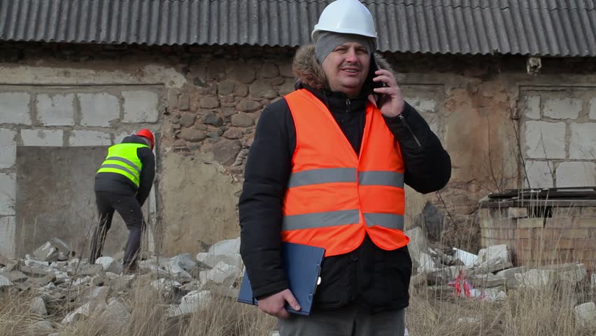 Building inspector talking on smart phone in background worker using slagdehammer