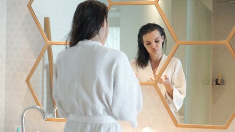 Young, pretty woman in bathrobe applying cream on face and neck in bathroom 
