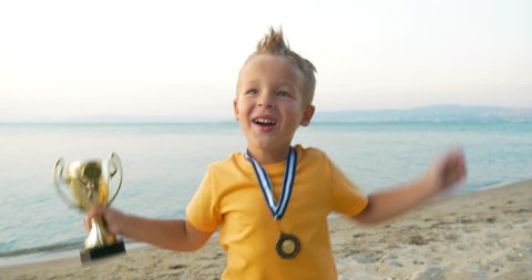 Little child with cup and medal jumping for joy to be the winner. He shouting and kissing prize at the seaside
