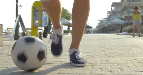 Steadicam close-up shot of man dribbling a soccer ball on the paved sidewalk in resort town, street cafes and hotels in background Video Stok