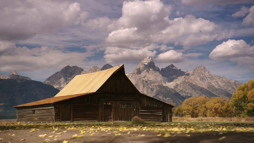 Beautiful classic shot of Teton mountain peaks and historic Mormon pioneer barn with autumn leaves blowing across is slow motion.  Dramatic clouds and sky. Grand Teton National Park, Wyoming, USA. 4K.