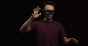 Young man wearing futuristic virtual reality headset scrolls and turns imaginary dials against black background, working or playing immersed in digital world