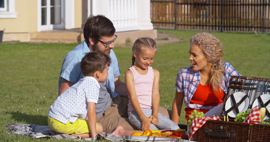 Happy family having picnic on green lawn: mother, father and little boy kissing cute daughter together and smiling