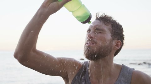 Fitness man drinking water from bottle splashing water in face cooling down after running workout on beach. Thirsty athlete having cold refreshment drink sweating after intense exercise. SLOW MOTION.