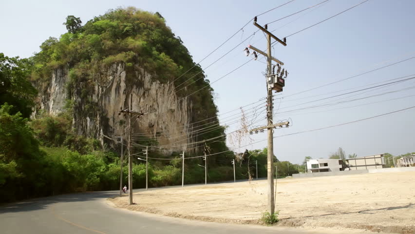 Motorcyclist going around road bend with over head power lines, empty cleared building land and karst limestone foliage covered hill with blue sky in background, Krabi Thailand