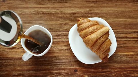 Pouring coffee in white cup and fresh baked croissant on wooden background. Top View.