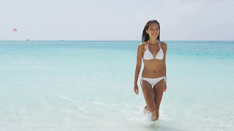 Sexy Asian bikini woman relaxing on beach walking out of turquoise ocean water wearing white fashion swimwear. Skin and hair care female model with slim body for weight loss or vacation concept.