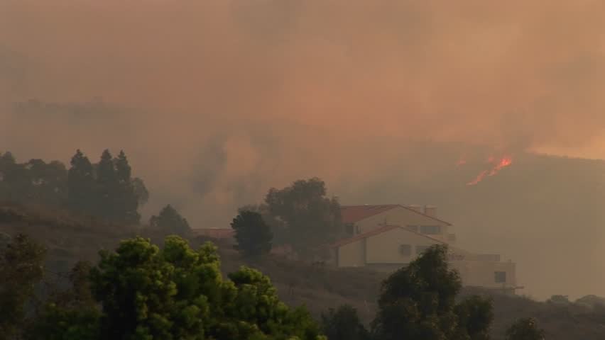 Long shot of a helicopter dropping chemicals over fires raging around a Southern California community