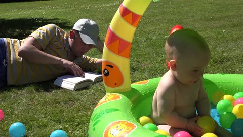 Serious student man dad study book and look after infant baby child sitting in kiddie pool full of colorful balls on garden meadow. Static shot. 4K