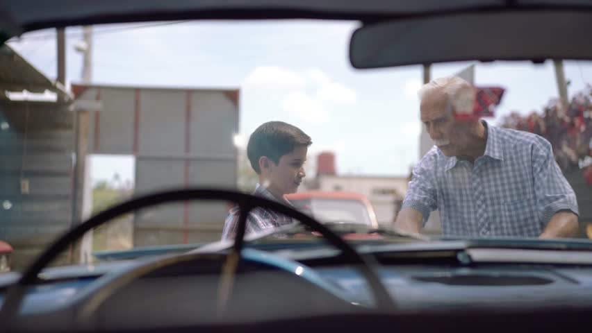Preteen child looking at the engine of a vintage car from the 50s. He is amazed and excited. The boy smiles happy and leans against the open hood. Viewed from the interior of the car
