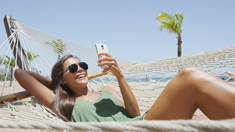 Happy beach woman reading sms texting on smartphone lying on hammock relaxing on tropical vacation. Young casual cute girl lying down in outdoor swing bed enjoying sun sunbathing using mobile phone.