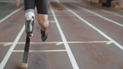 Tilt up of inspiring amputee athlete with artificial leg running on track at indoor stadium in slow motion