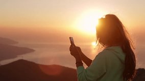 Young Attractive Woman Watching Sunset or Sunrise Over Bay from Top of the Mountain, Girl Takes Panorama Photos On Phone.Hair in the Wind. Handheld Atmospheric Shot. Crimea, Ukraine. 