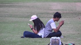 Young Asian couple relaxing in the park and playing with smart phones and taking selfies. Filmed in 4k video in Bangkok.