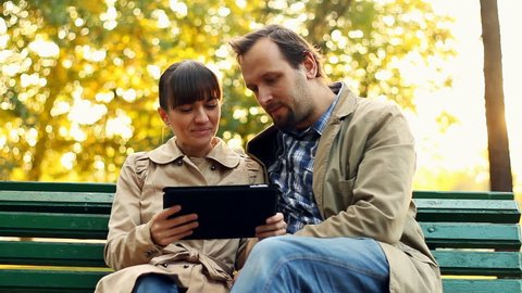 Young couple with tablet computer sitting on bench in autumn park