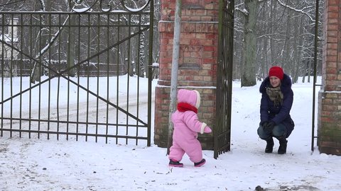 Happy mother with baby daughter girl walk through retro gate in snowy winter park. Static shot. 4K