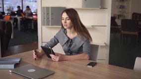 Female sitting at the personal office on shelves documents in  folders. Woman speaking on touch screen tablet. She using earphones for speaking. They discussing project. Lady with long hair smile and
