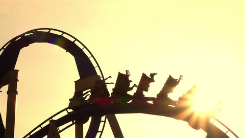 SLOW MOTION CLOSE UP: People riding extreme roller coaster attraction in amusement park at golden sunset. Rail looping coaster ride over the setting sun.