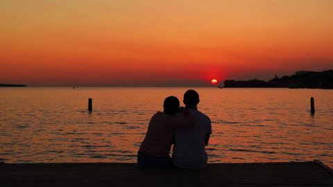 Romantic family scene: silhouette of couple at the sea background. Family couple sitting on the beach. The couple looks at the sunset on the Adriatic sea. A pair of lovers watching the sunset. Piran
