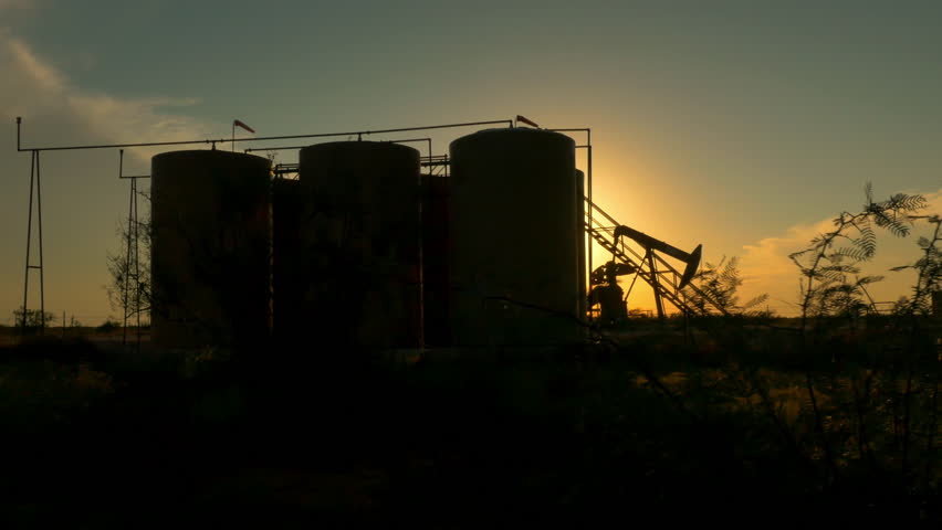 CLOSE UP: Industrial oil pump jack pumping crude oil and storing it in big reservoir containers in oil field. Nodding donkey pump working over the setting sun in the middle of desert at golden sunset
