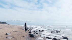 Woman on the cold icy deserted beach taking photo or video of ice breaking and sea. Blues skies, ice, ice floes, open water, clouds and horizon,