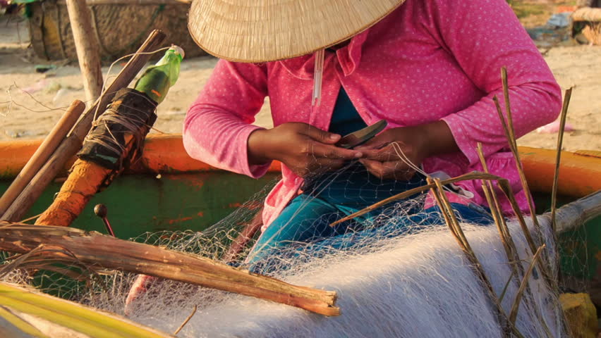 closeup vietnamese woman in hat sits in round boat and mends fishing nets on sand beach