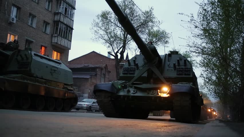 Tanks with big towers and armored vehicles with soldiers riding on street of Samara near houses and shops on military celebration.