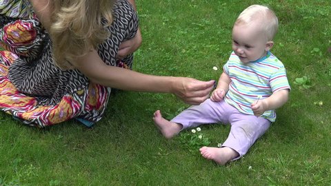 Mother pick daisy flower and give it to cute baby exploring nature siting on lawn. Woman share knowledge with interested infant child finding new discoveries. Static closeup shot.