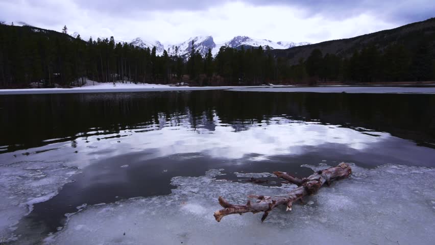 Melting lake at the base of a forest and snow capped mountains.