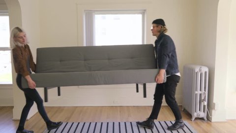 Young Trendy Couple Move A Couch Into New Apartment, They Set It Down, Adjust It, Then Sit And Kiss