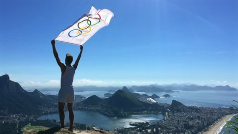RIO DE JANEIRO - MARCH 21, 2016: Athlete stands holding Olympic flag above a city skyline view of Corcovado Mountain and Zona Sul. Editorial Stock Video