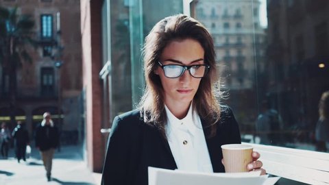 Close-up of young attractive businesswoman wearing glasses and drinking coffee to take away while working with documents outdoors, looking serious and concentrated, slow motion