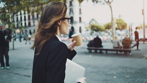 Close-up of young professional businesswoman walking outdoors, drinking coffee to go and holding modern portable computer or laptop, slow motion