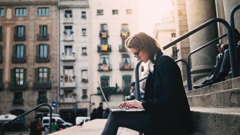 Close-up of young attractive businesswoman using laptop on a break outdoors, beautiful female student sitting on the stairs of University, slow motion