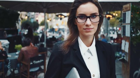 Close-up portrait of young beautiful businesswoman looking at camera with smile outdoors, professional female cafeteria manager wearing glasses and black suit, slow motion
