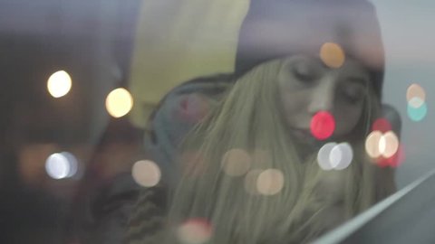 Teenage blonde girl in a beanie and scarf waking up and falling asleep in a public transport next to window with city lights reflection on the window. Exhausted, tired, sleepy woman in a train. 