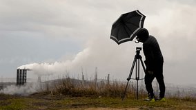 Man Photographer Closes the Camera on a Tripod Umbrella From the Rain and Wind. Survey of Industrial Enterprise.