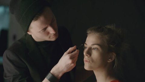 Young male makeup artist brushing eyebrows of a fashion model while preparing her for photo shoot