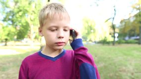 Little child with cell phone close to ear walking in urban park. Real time video footage of 8 years old beautiful blond boy outside on summer or early fall warm day.