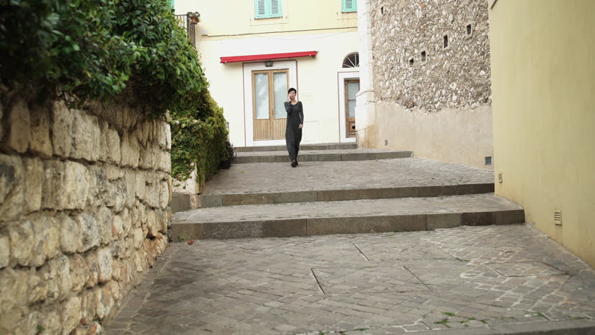 Girl walking on stairs on the street in old city. Female using app on mobile phone. She dressed in casual long grey dress. Mixed race model happy and smile. Tourist wander in mediterranean city in