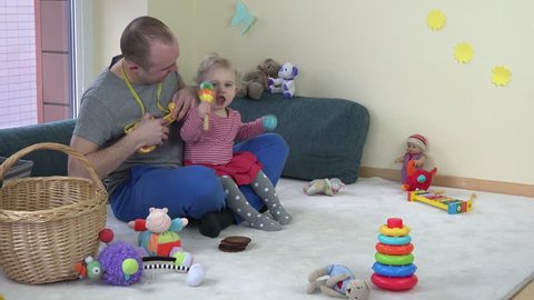 Man play toy guitar and toddler girl dance with rattles. Family have fun at home. Static shot.
