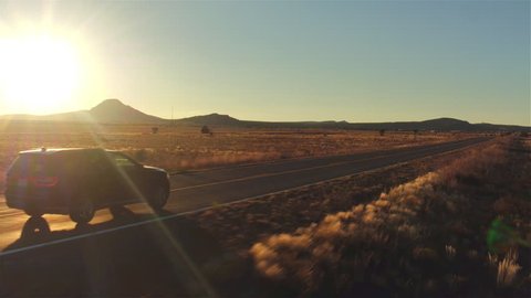 AERIAL: Black SUV car driving along empty country road at golden summer sunset. People traveling, road trip on historic Route 66 through beautiful meadow desert scenery in sunny summer