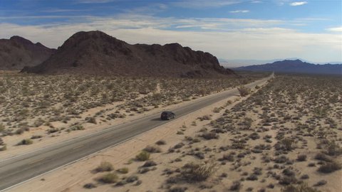 AERIAL: Black SUV car driving along the picturesque empty road through the vast desert with big rocky mountains. People traveling, road trip through beautiful countryside scenery in sunny summer