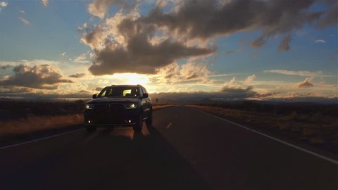AERIAL: Black SUV car driving along the picturesque empty road through the vast desert at beautiful golden sunset. People traveling, road trip through beautiful countryside scenery in sunny summer