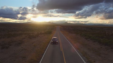 AERIAL: Black SUV car driving along the picturesque empty road through the vast desert at beautiful golden sunset. People traveling, road trip through beautiful countryside scenery in sunny summer