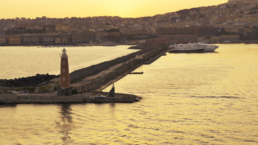 POV from passing cruise ship. Golden evening lights the sky and water leaving Naples, Italy and passing the lighthouse at the end of the jetty and houses and buildings along the shoreline.