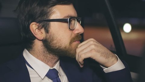 Close up of tired businessman sitting in the back seat of taxi cab, looking in the window then taking off his eyeglasses and rubbing chin