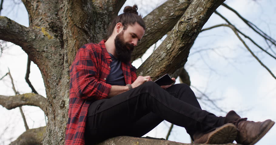 4k young carefree man sitting on a tree trunk away from hustle and bustle of a city and browsing the internet on a touchscreen tablet..