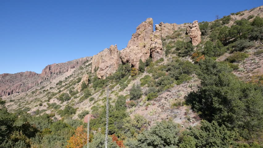 A short pan across tall rock formations on a hill with shrubs and cedars in the Chisos Mountains of Big Bend National Park.