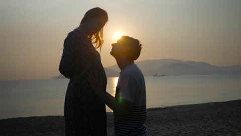 Pregnant woman and man on bended knees caressing and kissing his wife's belly on a sea sunset background.